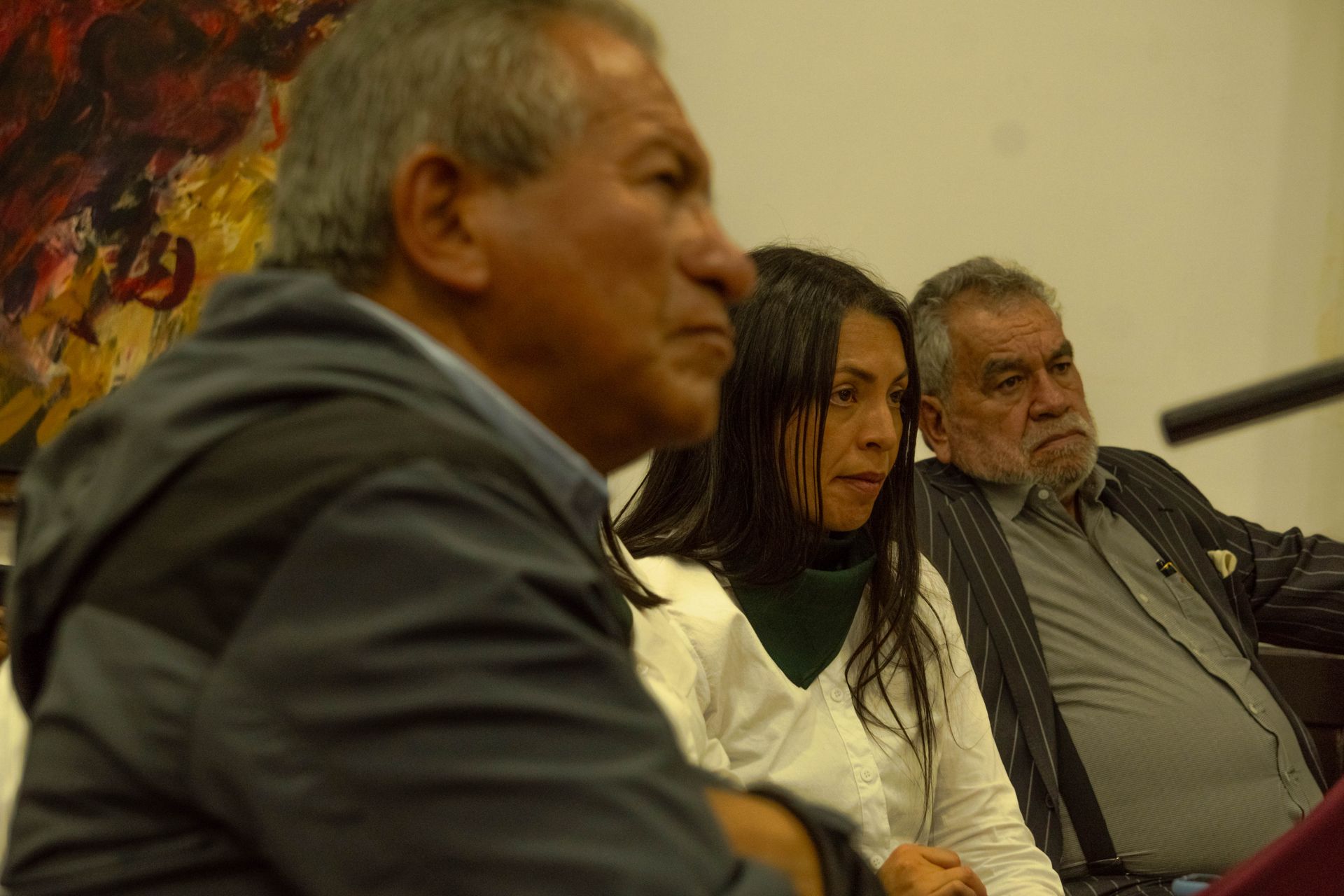 Three people sit indoors, focused. Man in front has gray hair, woman in white looks intently, older man on the right with a beard.