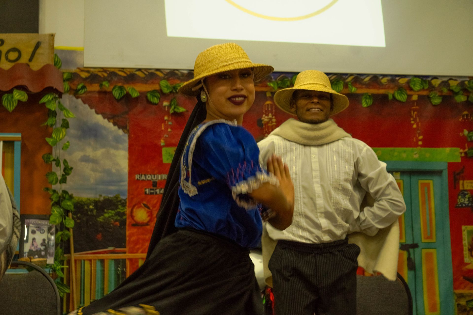 Two people in traditional attire dancing, smiling, in front of a colorful backdrop, wearing hats.