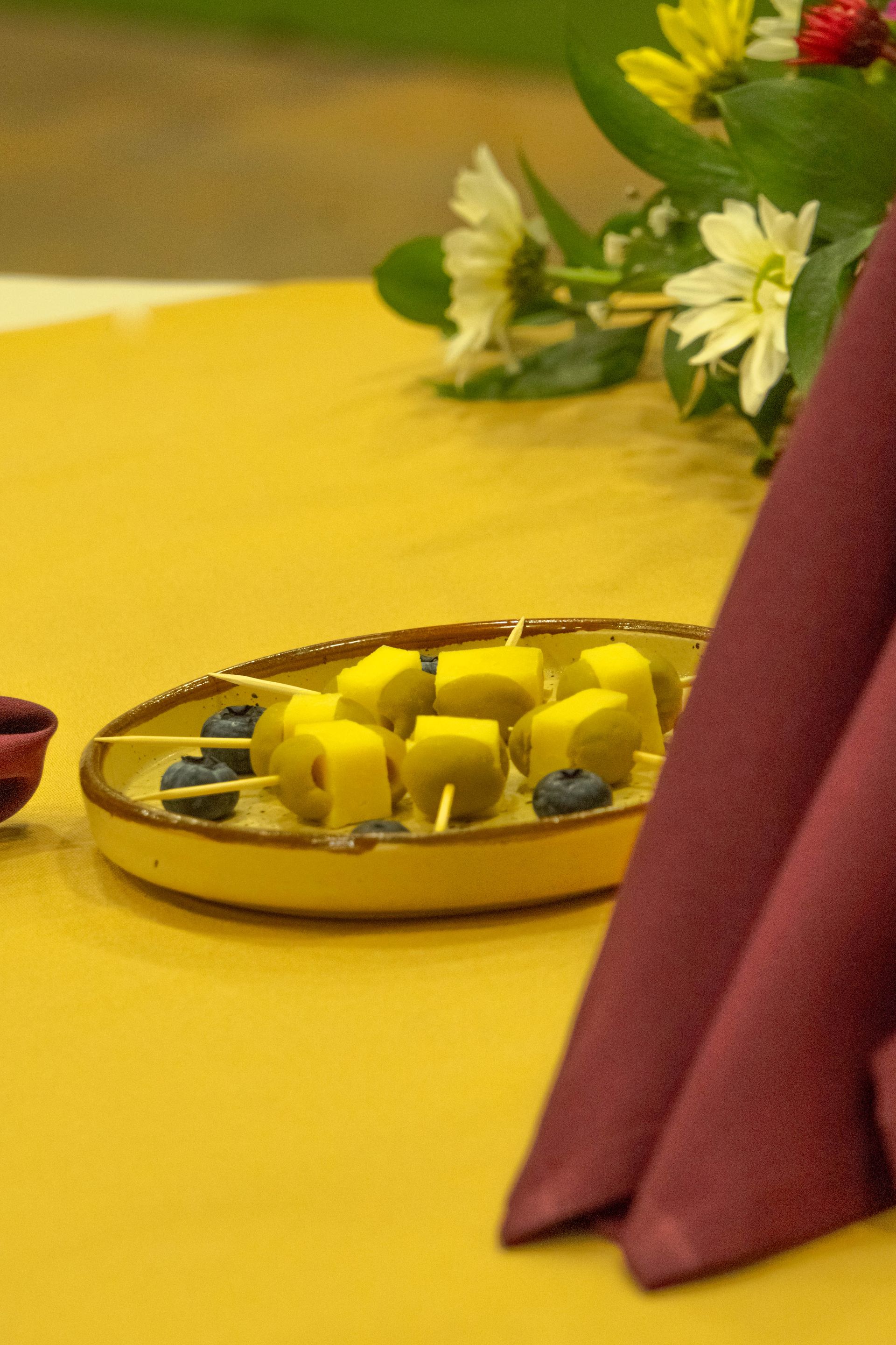 Yellow plate with mango skewers and blueberries on a yellow tablecloth, next to a red napkin and flowers.