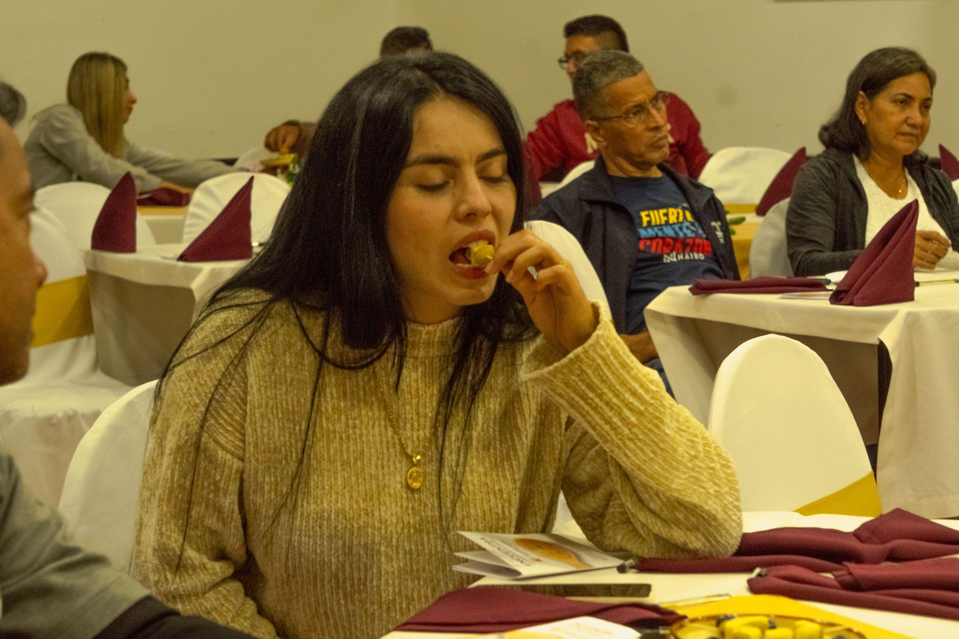 Woman eating food at a table, others seated in a banquet hall.