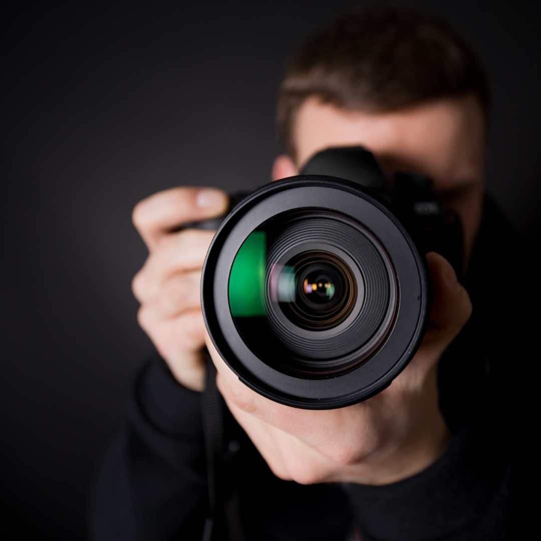 Person holding a camera, lens pointed forward, against a dark background.