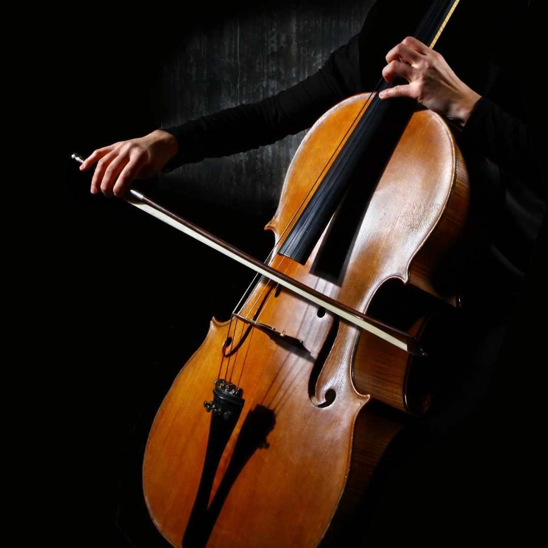 Person playing a cello, with hands on the bow and neck of the instrument; dark background.