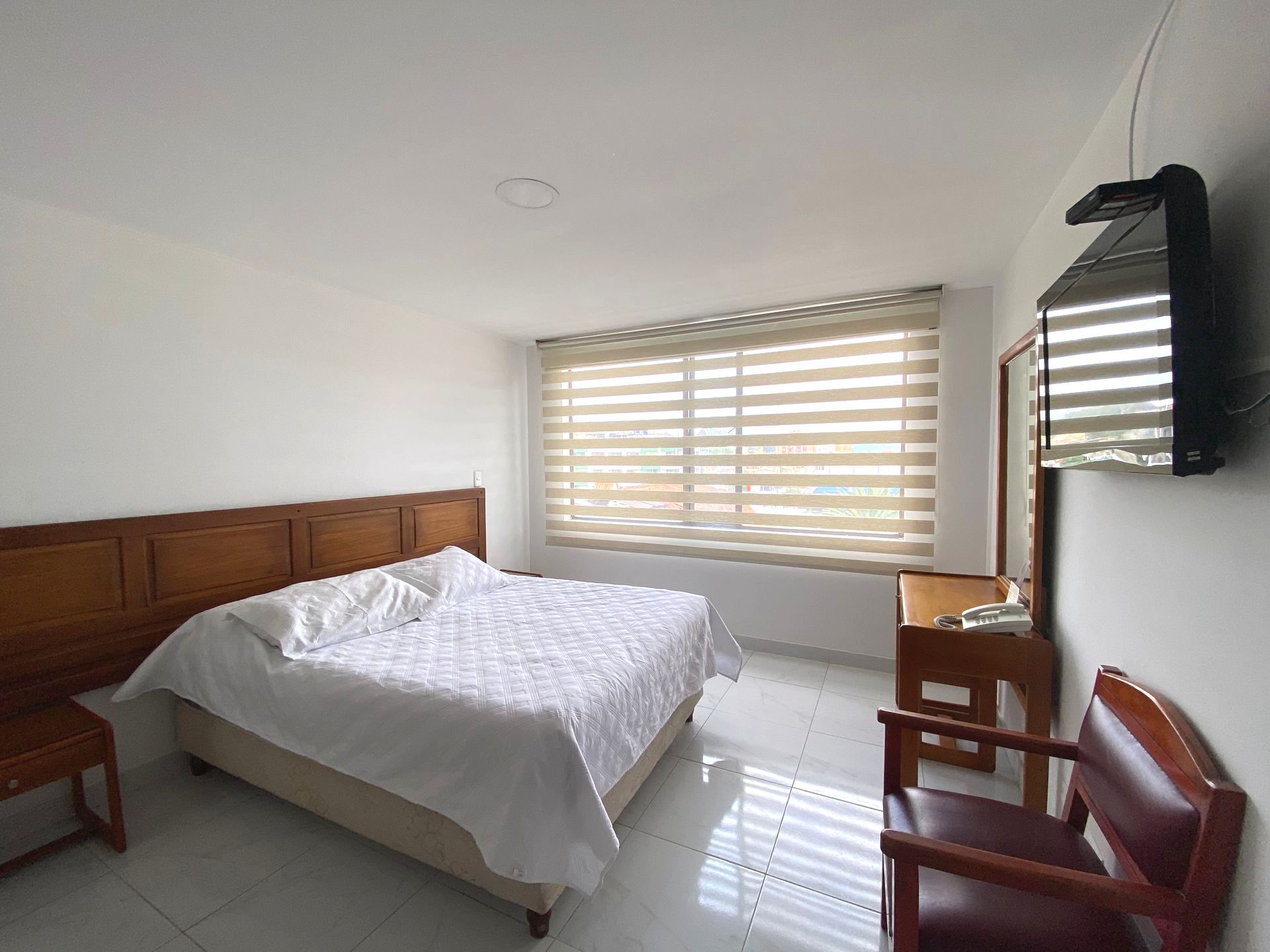 Bedroom with a bed, nightstand, chair, window with blinds, and a wall-mounted TV.