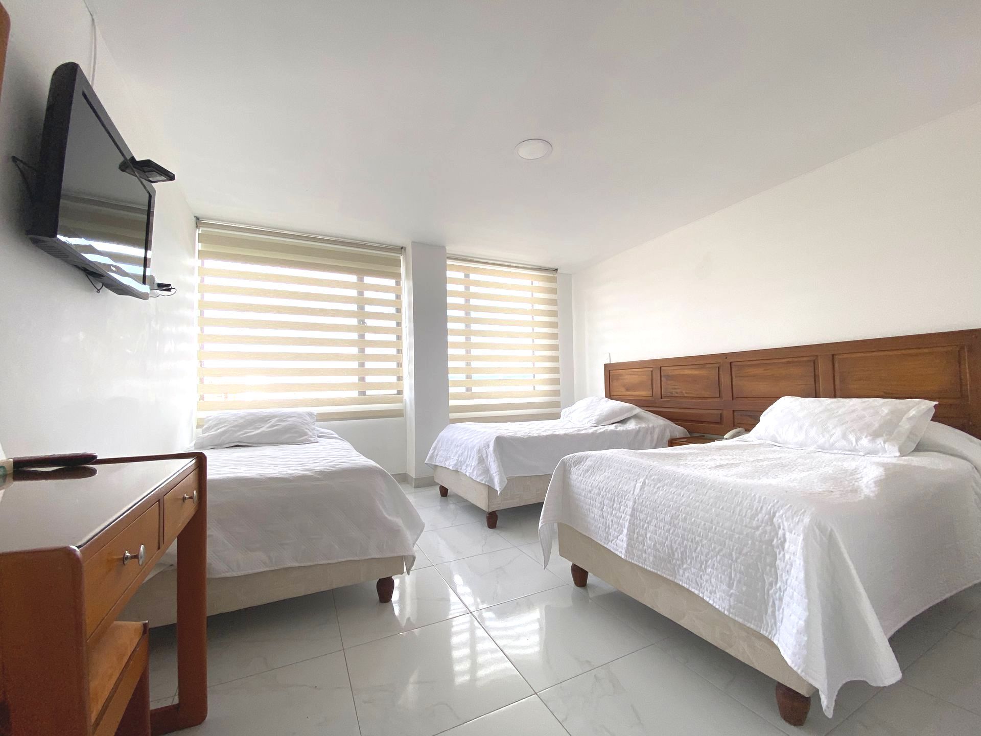 A hotel room with three single beds, a desk, and a mounted TV. White walls and bright lighting.