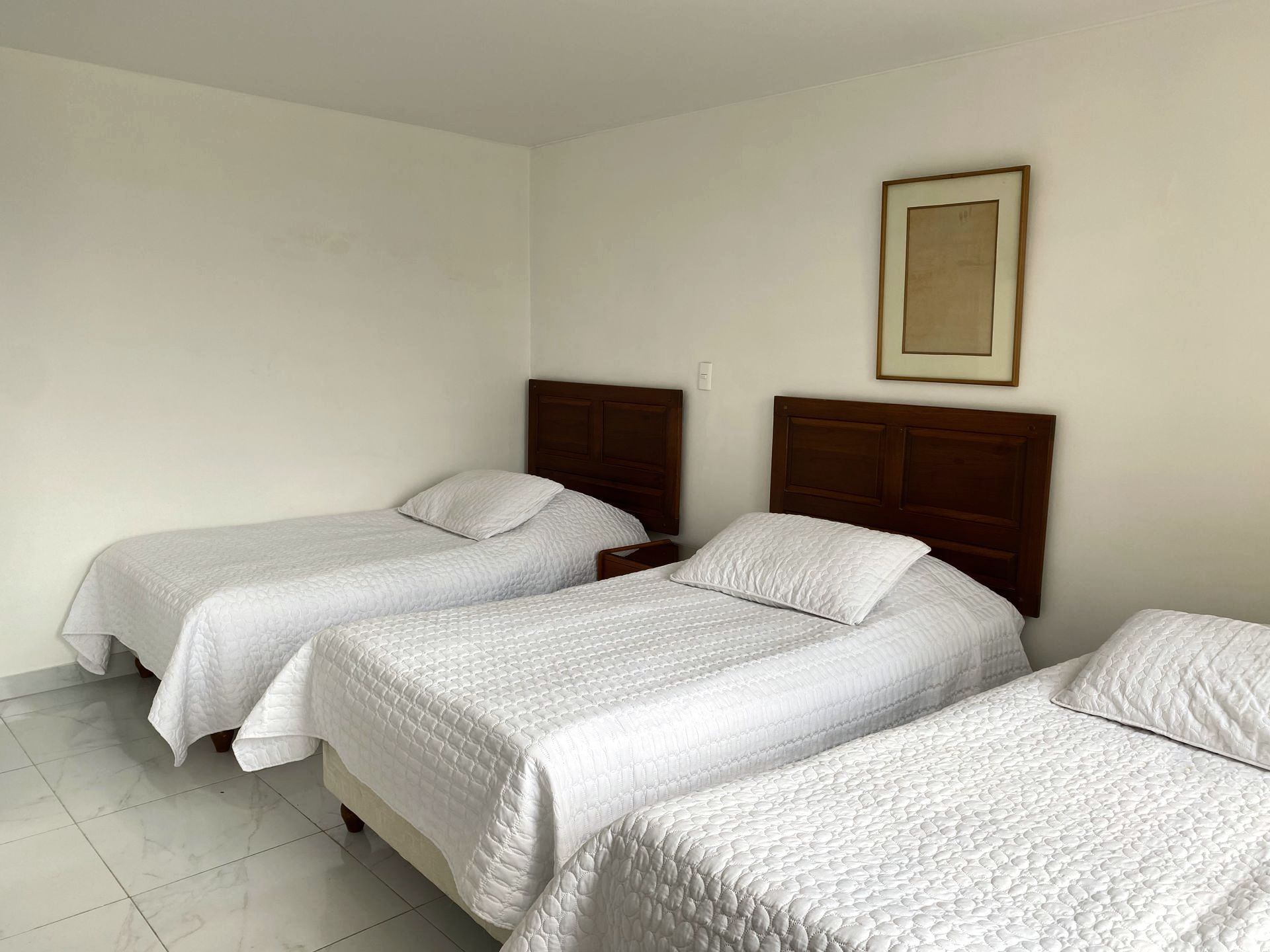 Three twin beds in a plain white room. One framed artwork hangs above a bedside table.