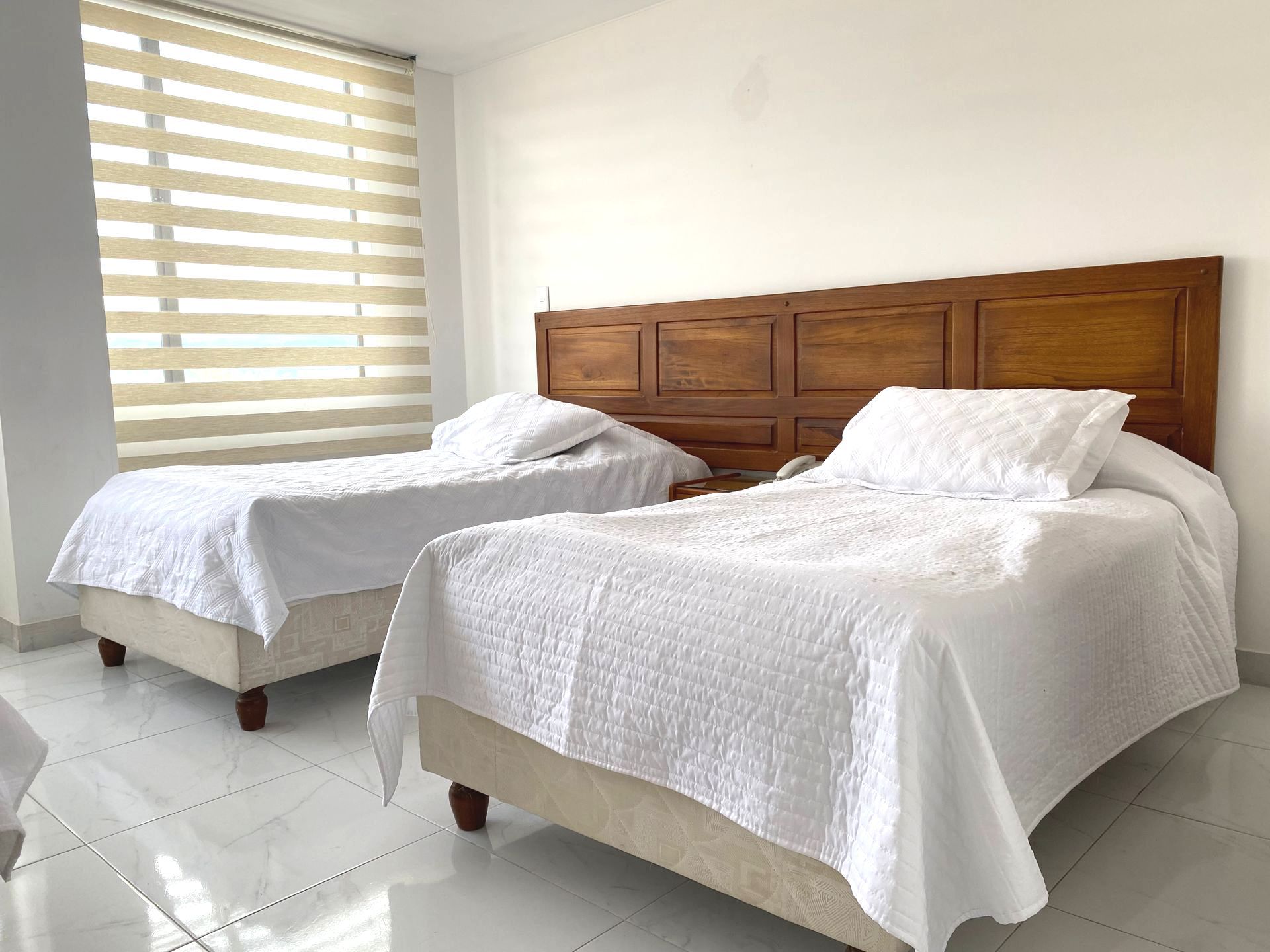 Two twin beds with white bedding, in a bright room with a wooden headboard and window blinds.