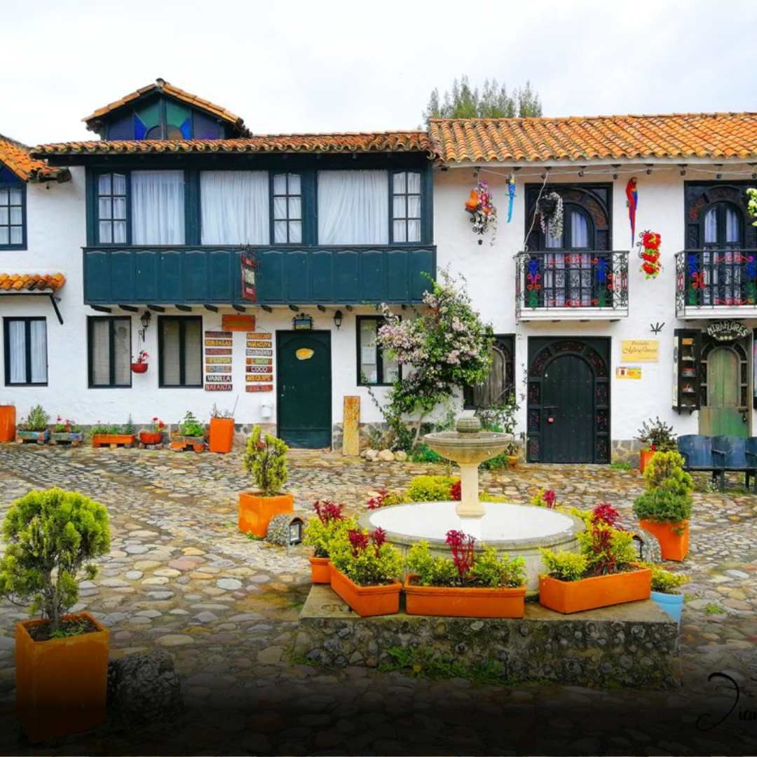 Courtyard with white buildings, balconies, and a fountain, surrounded by flower pots, in a cobblestone square.
