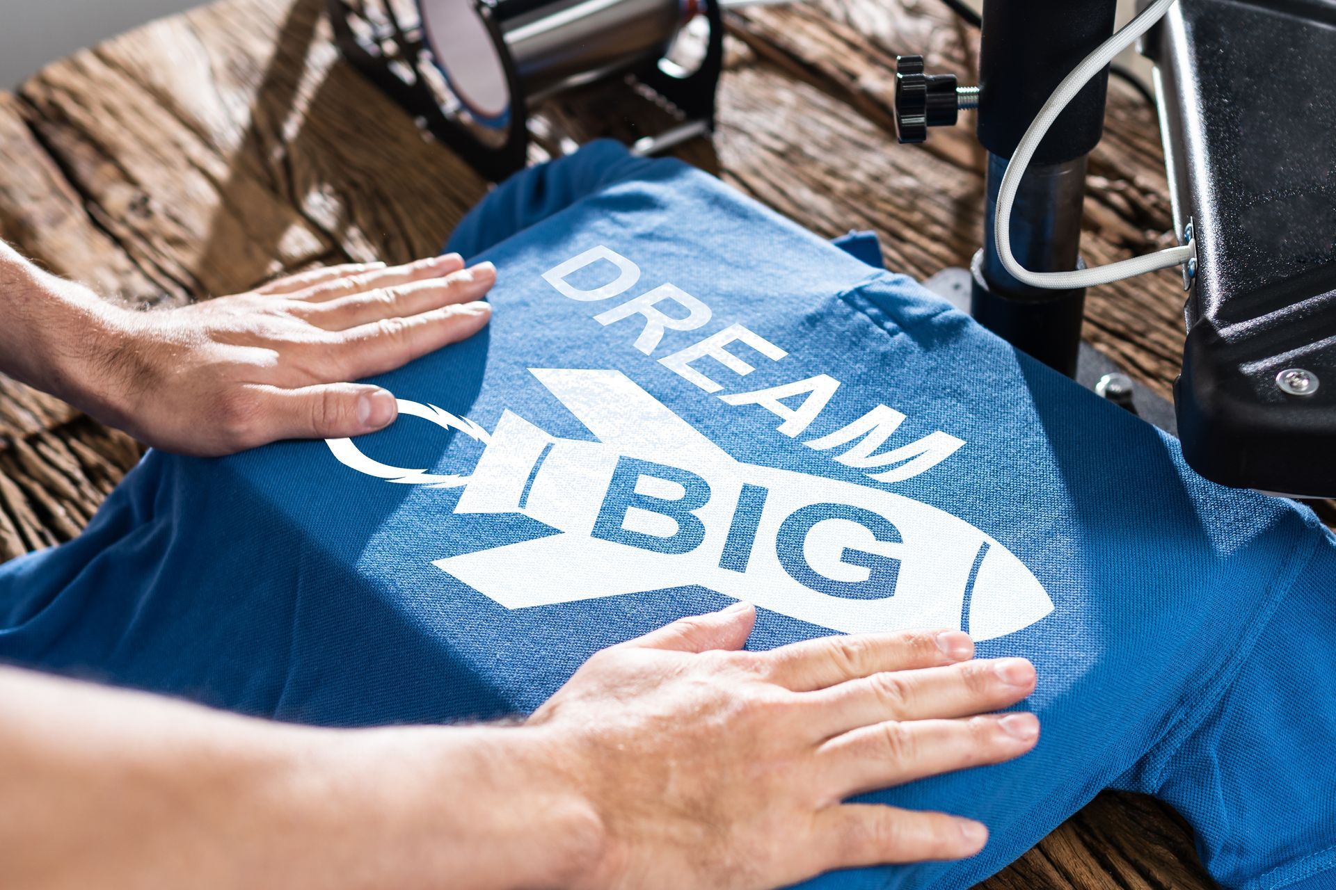 Hands smoothing a blue T-shirt with a white “DREAM BIG” rocket design under a heat press machine on a wooden table.