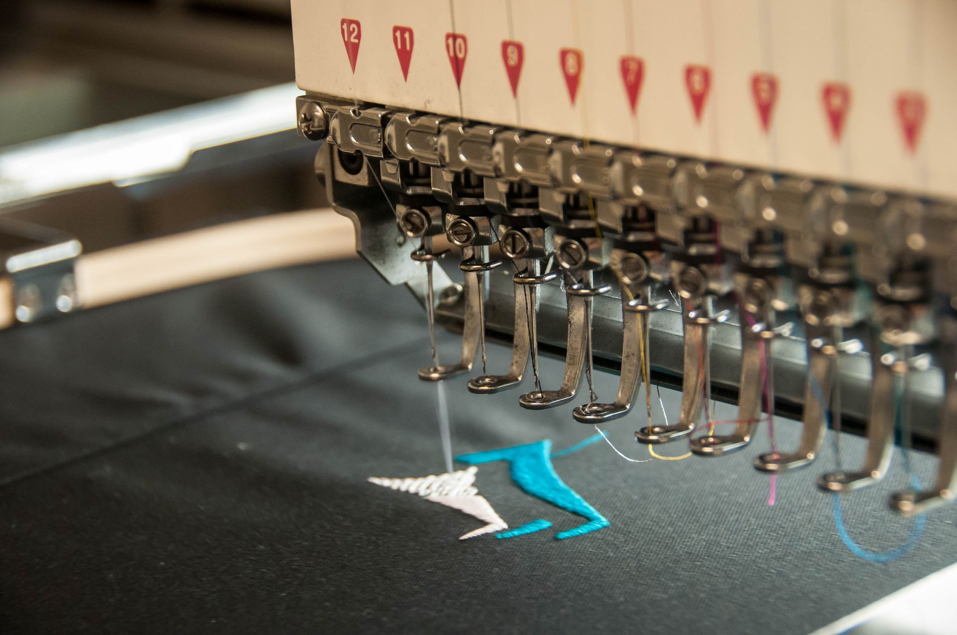 An industrial embroidery machine stitching white and turquoise thread patterns onto black fabric.
