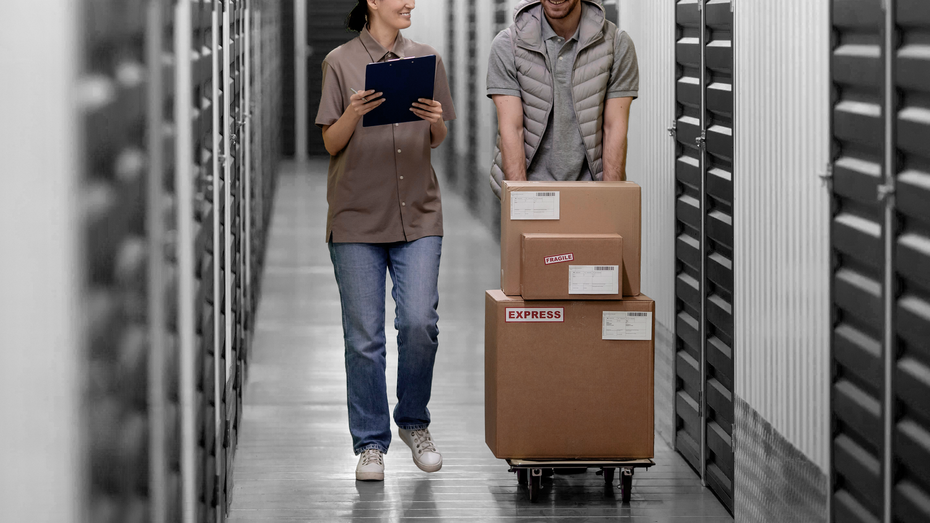 A man and a woman are carrying boxes on a cart in a warehouse.