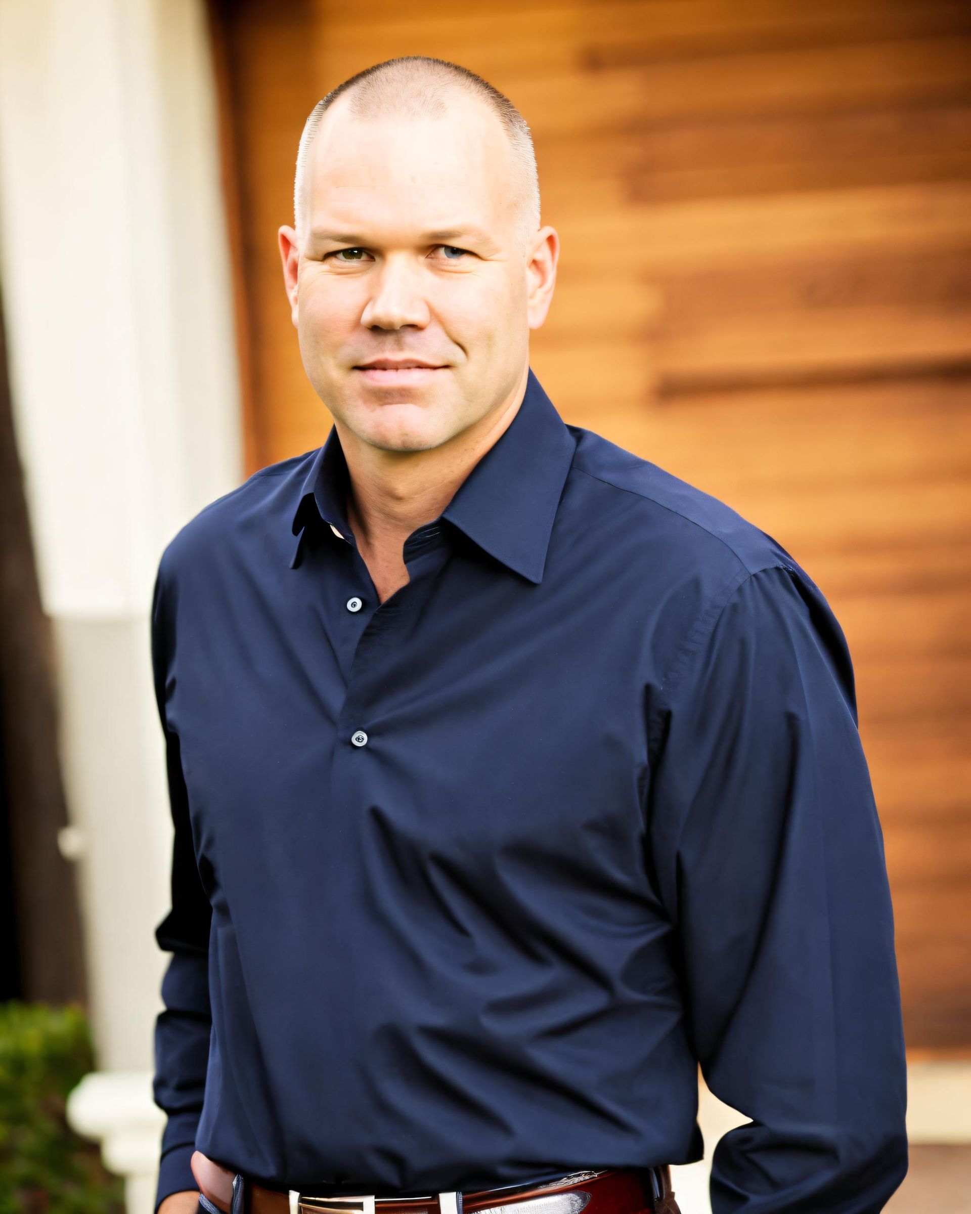 Man with shaved head in blue shirt, smiling slightly, outside by wooden paneling.
