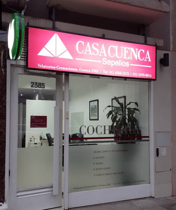 Fachada de funeraria con cartel iluminado en rojo, puertas de vidrio y dirección 2385.