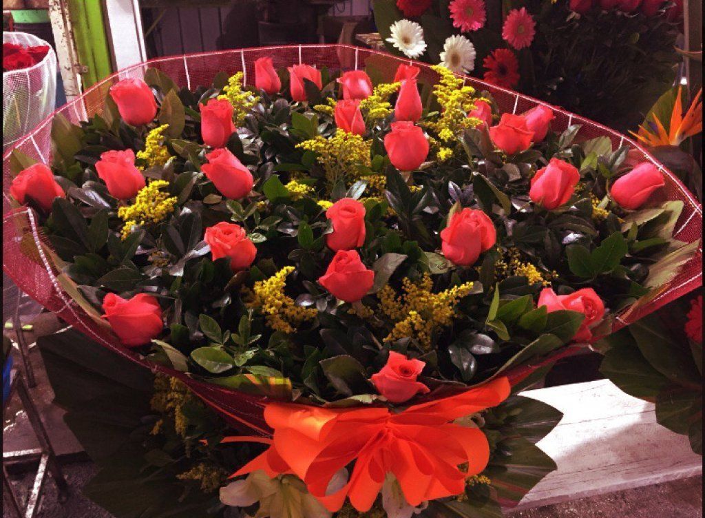 Ramo de rosas rojas vibrantes y flores de relleno amarillas, envuelto en papel de seda rojo con un lazo naranja brillante.