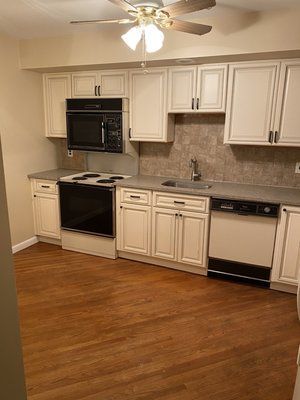 A kitchen with white cabinets , a stove , a microwave , a sink and a ceiling fan.