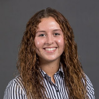 Smiling person with curly hair in a striped shirt against a gray background