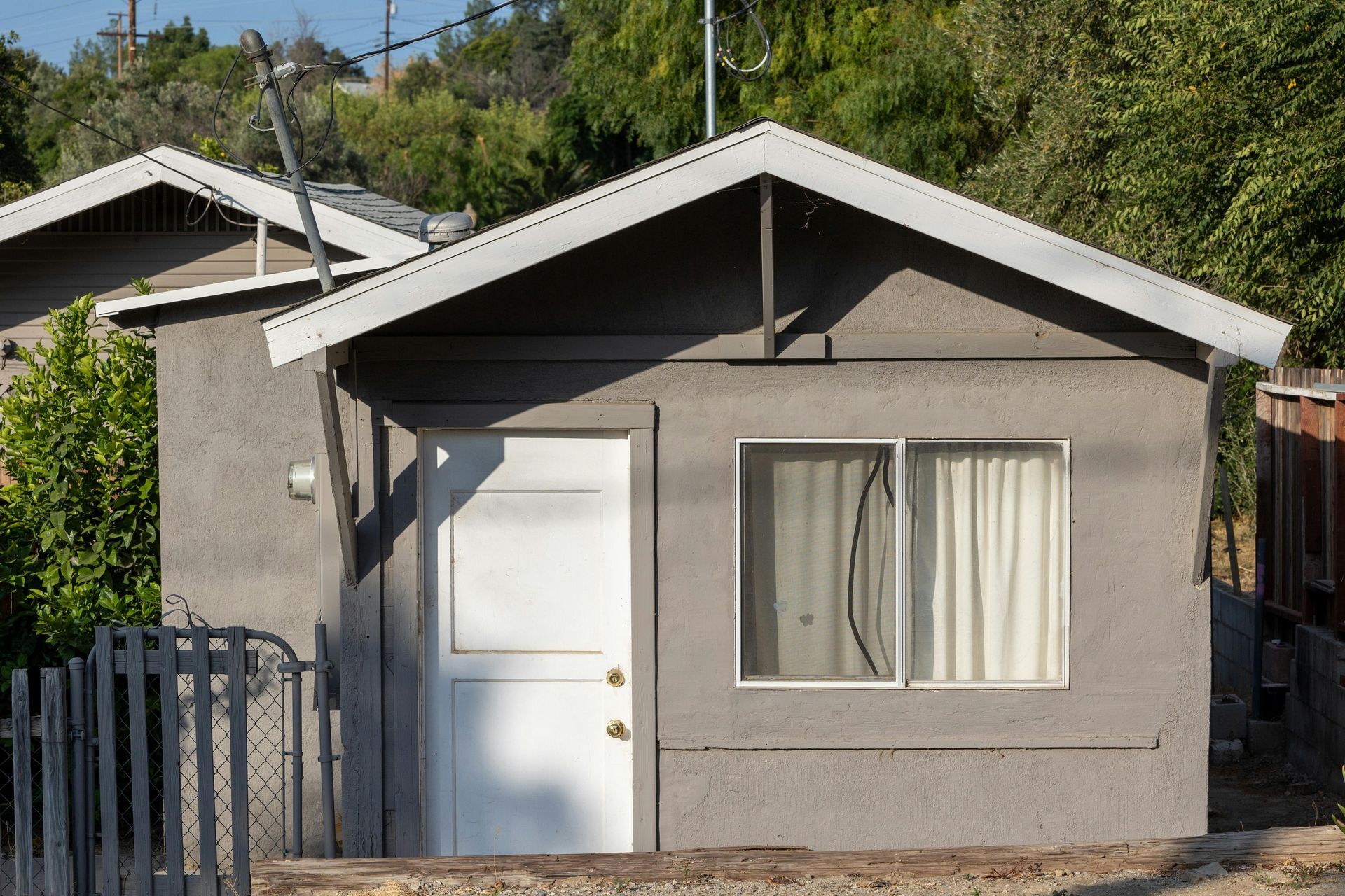 Small gray house with a white door and window, viewed from the front in a sunny yard.