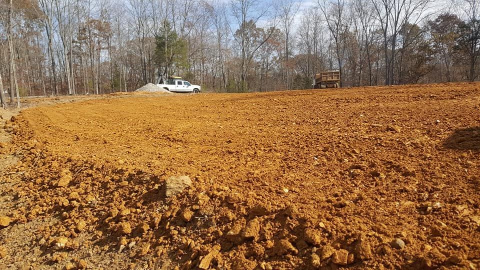 Brown, freshly graded earth; white truck and equipment in background; trees, open area.