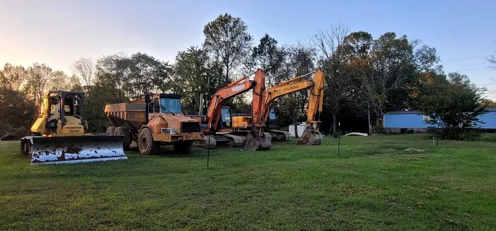 Construction equipment on a grassy field; includes a bulldozer and two excavators. Trees in the background.