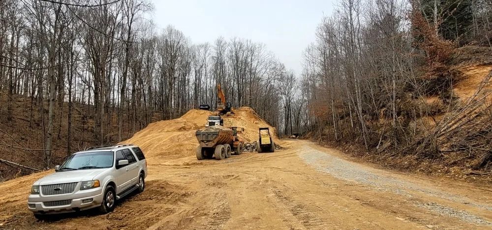 Construction site with heavy machinery, a vehicle, and a gravel road surrounded by trees.