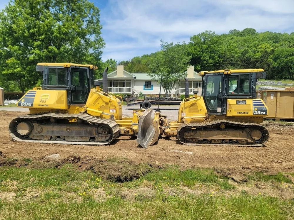 Two yellow bulldozers side-by-side on a construction site, with a house and trees in the background.