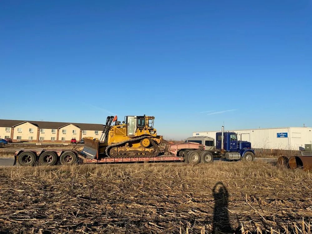 Yellow bulldozer on a flatbed trailer pulled by a blue semi-truck on a field, clear blue sky.