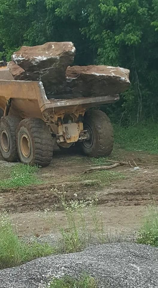 Dump truck carrying large rocks on a dirt road. Trees in the background.