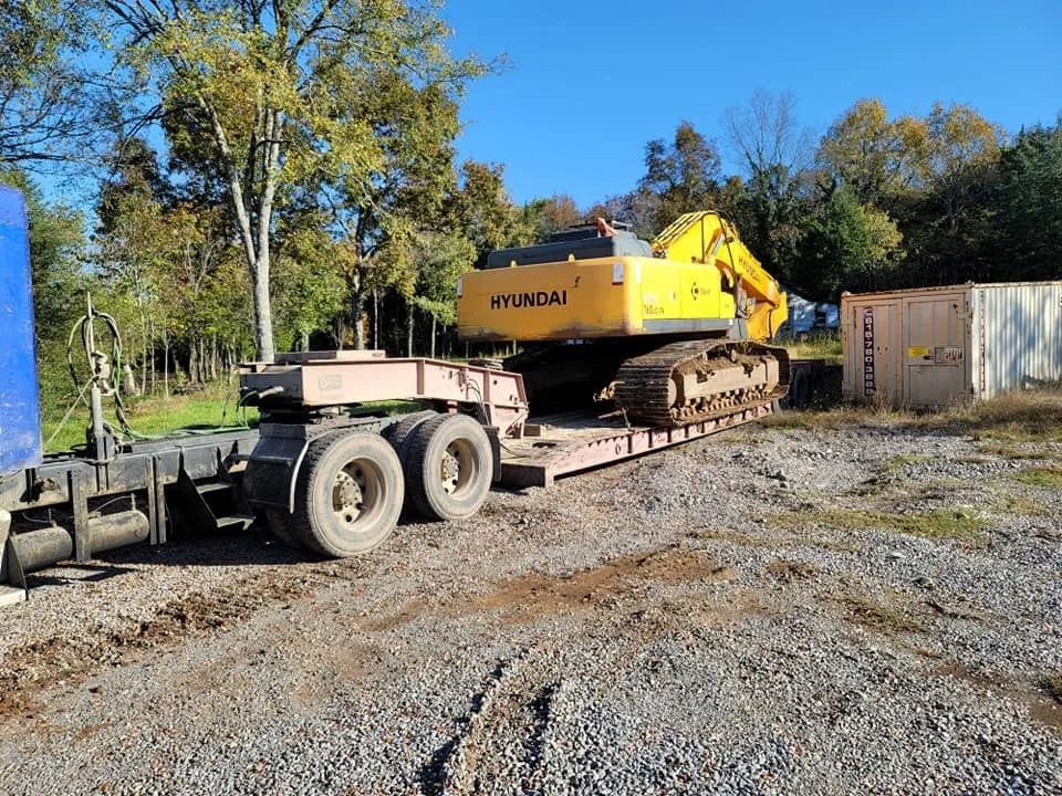 Yellow excavator on a trailer parked on a gravel lot near trees on a sunny day.