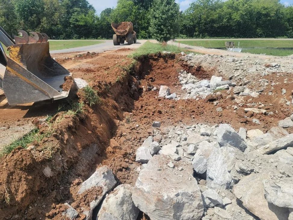 Excavator digging a trench next to broken concrete, in a park-like setting.