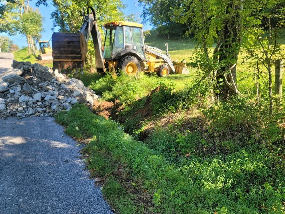 Yellow backhoe excavating a ditch next to a road, with a pile of rocks.