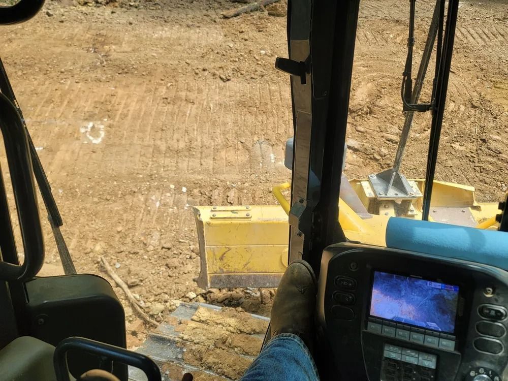 Inside bulldozer cab, view of worksite with yellow blade, foot on console, monitor screen visible.