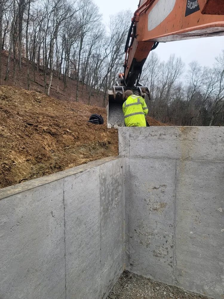 Construction site: excavator and worker in neon vest near a concrete retaining wall, hillside in background.