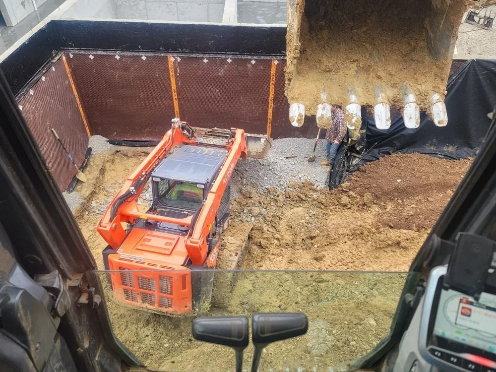 Excavator and skid steer working in a construction pit. Soil, black liner, and a person are visible.