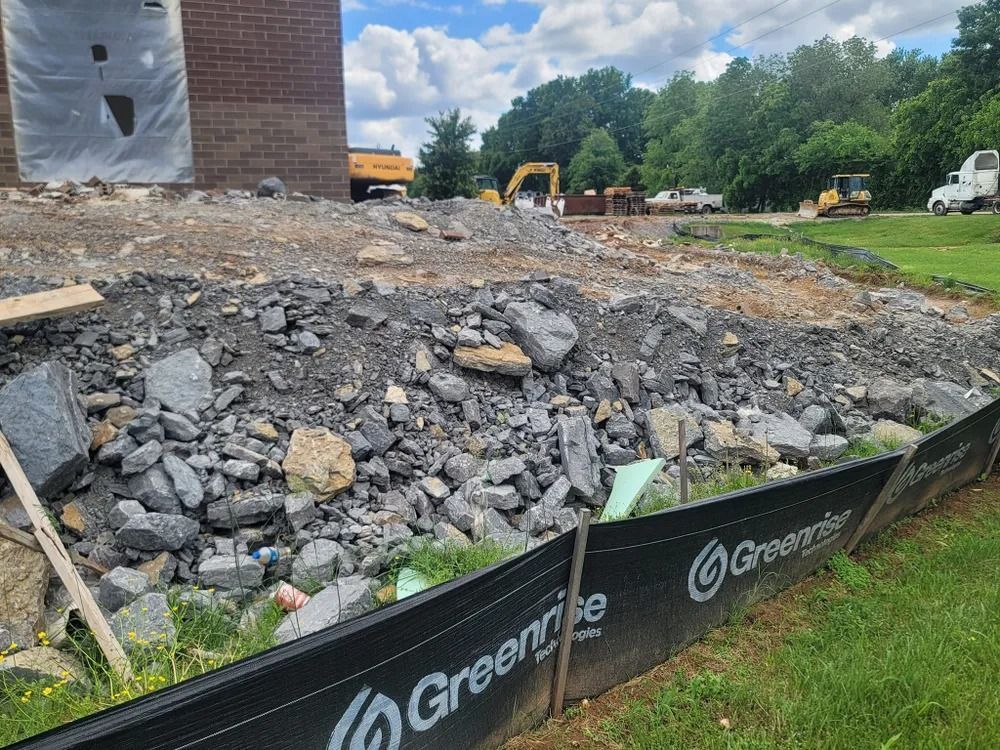 Construction site with a pile of rocks and dirt, surrounded by a black barrier, with heavy machinery in the background.