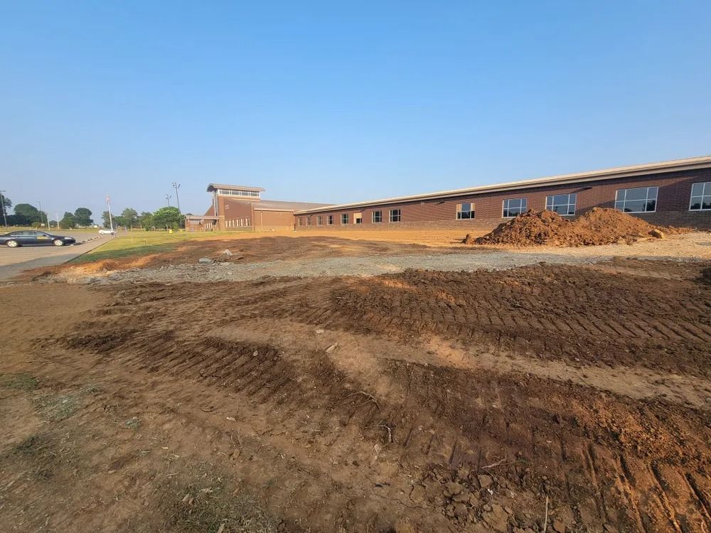 Construction site; a long brick building in the background. Brown earth and tire tracks in the foreground. Blue sky.
