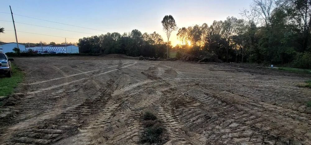 Cleared lot with tire tracks in the dirt, trees, and buildings in the background. Sunset.