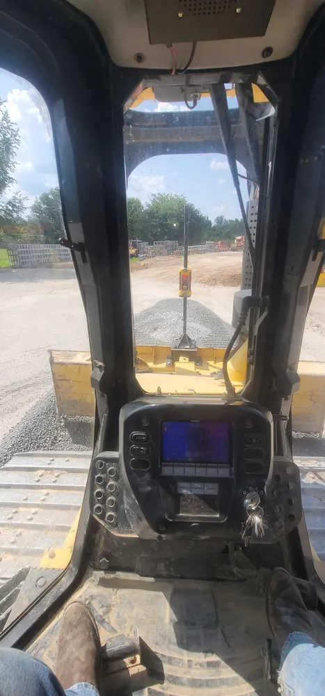 Inside view of a skid steer operator cabin, looking out at construction site. Yellow shovel is moving gravel.