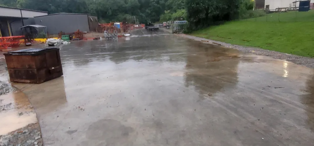 A wet, paved area reflects the overcast sky, with a dark, boxy object in the foreground and greenery in the background.