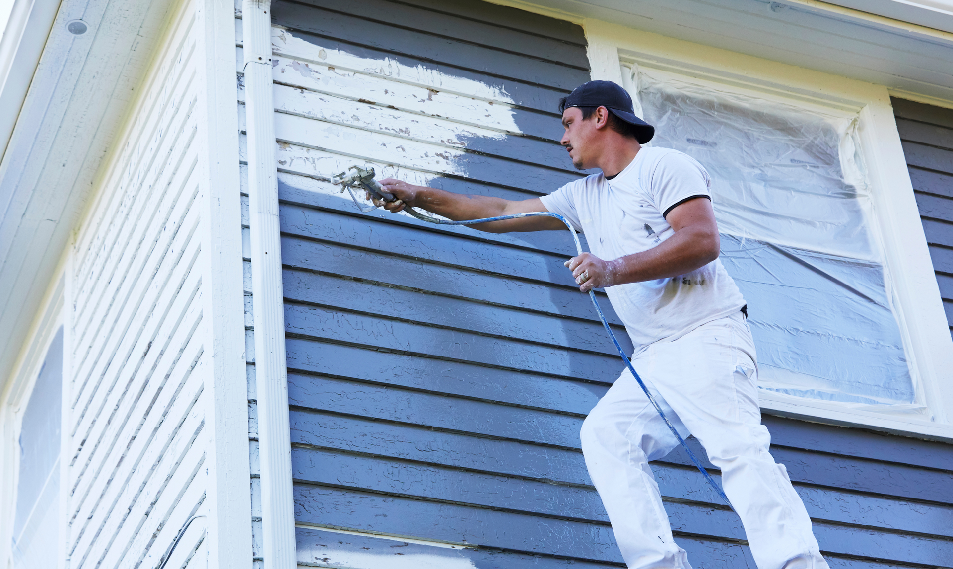 Man on a ladder painting house siding with a sprayer. White paint covering blue siding.