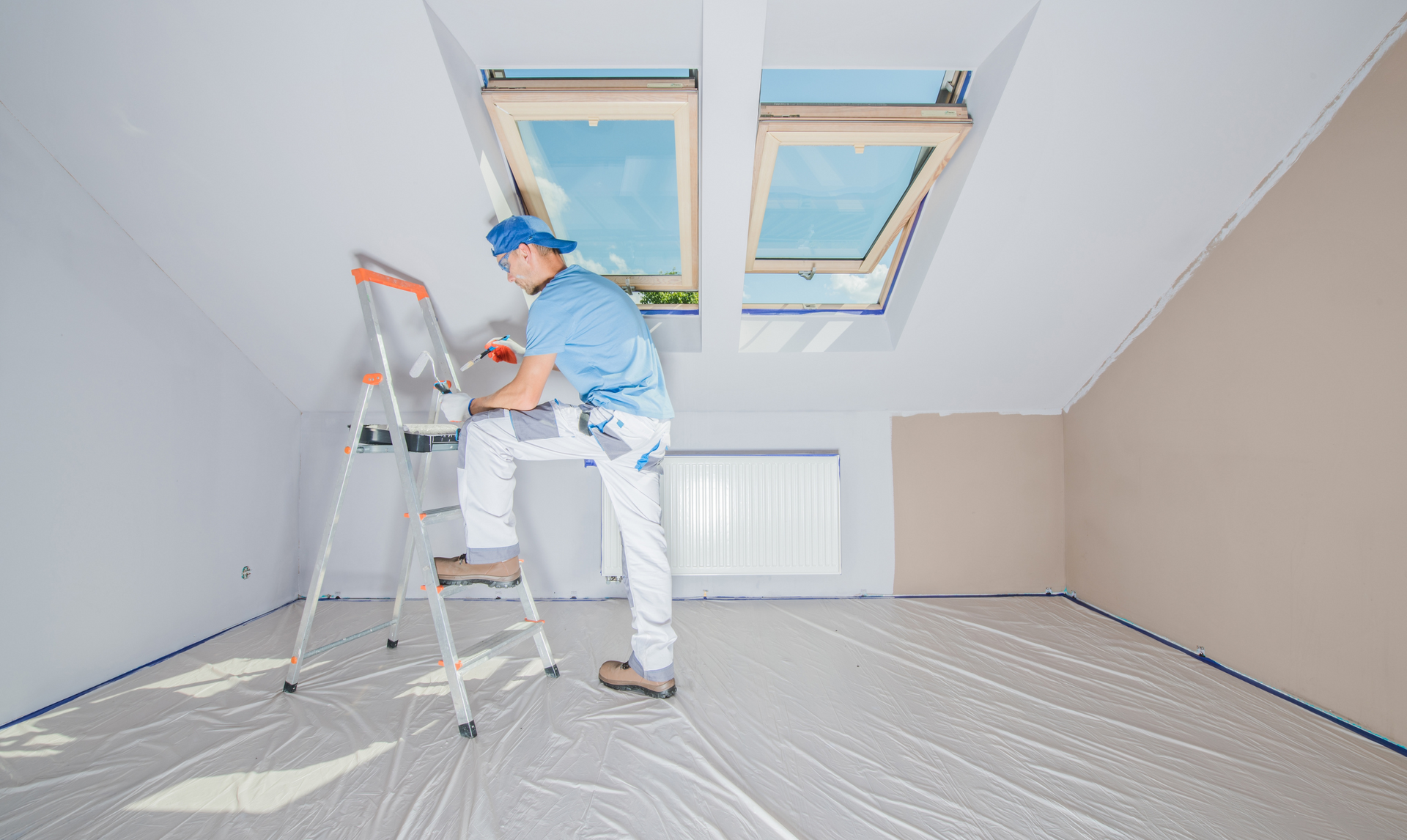 Painter on a ladder, painting a white ceiling with skylights in a room with tan walls, floor covered in protective plastic.