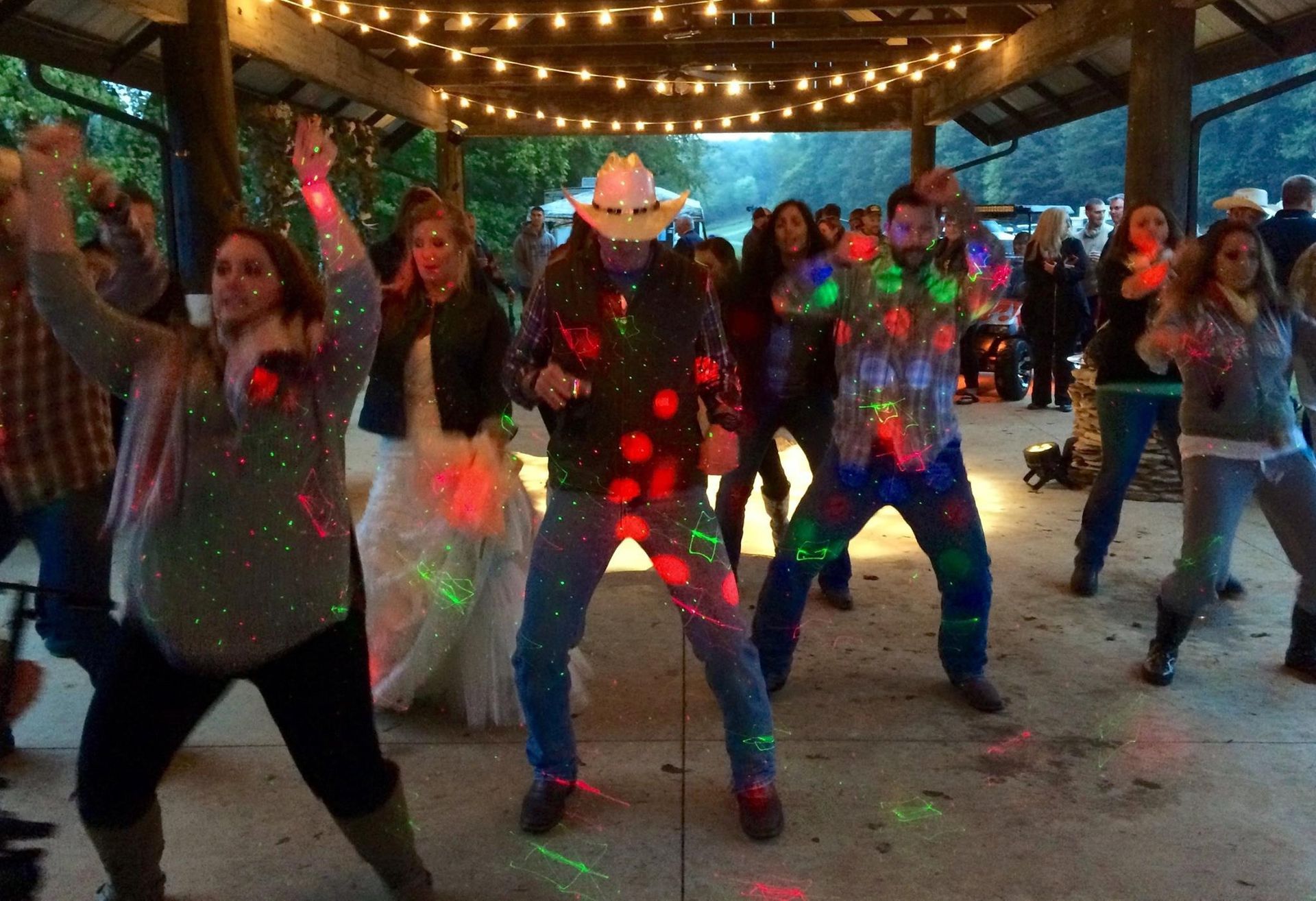 A group of people dancing under string lights at an outdoor pavilion, illuminated by colorful, moving disco laser lights.