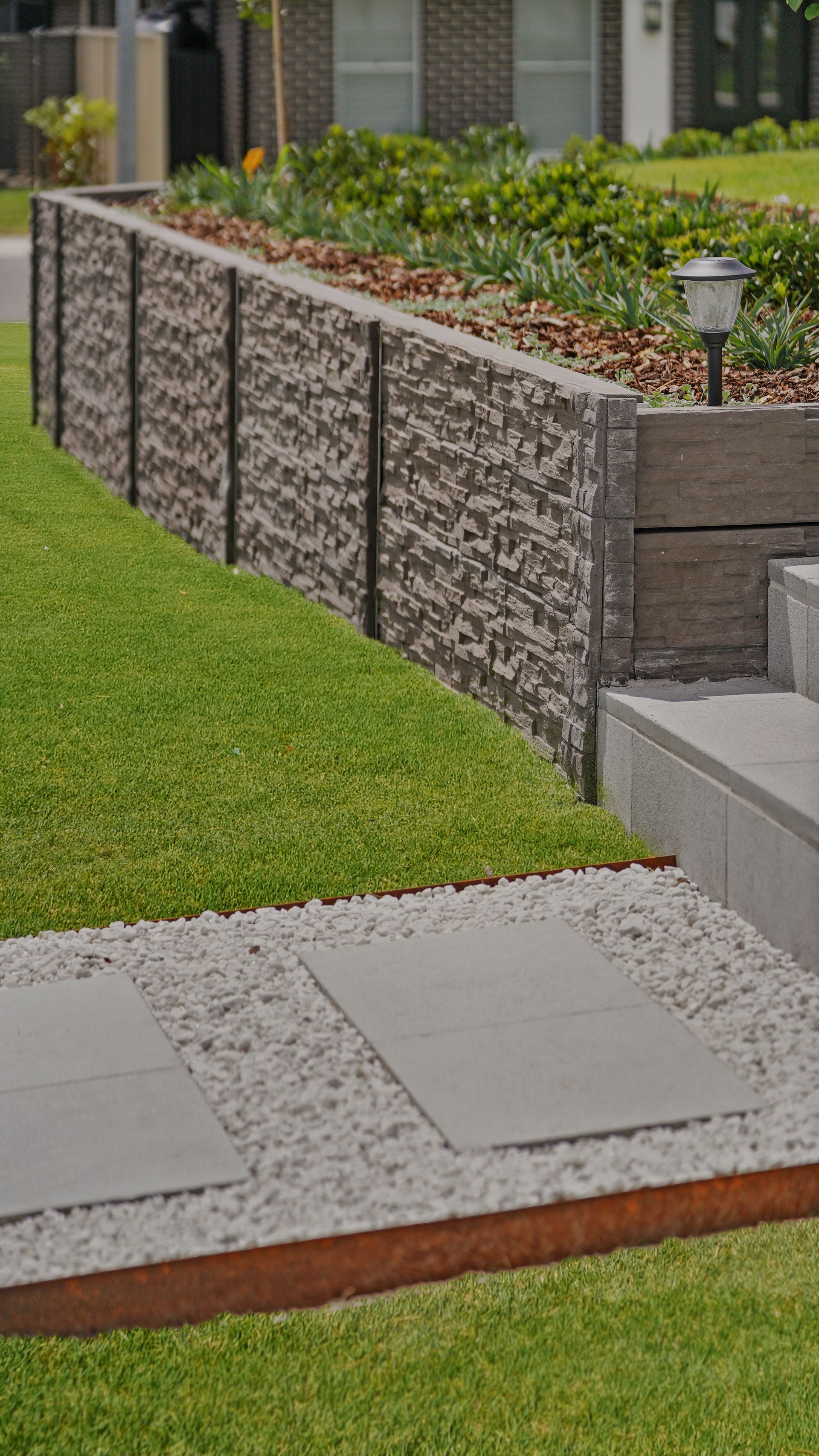 Modern landscape with concrete sleeper retaining wall, paving steppers in pebbles, and lush lawn in a residential yard