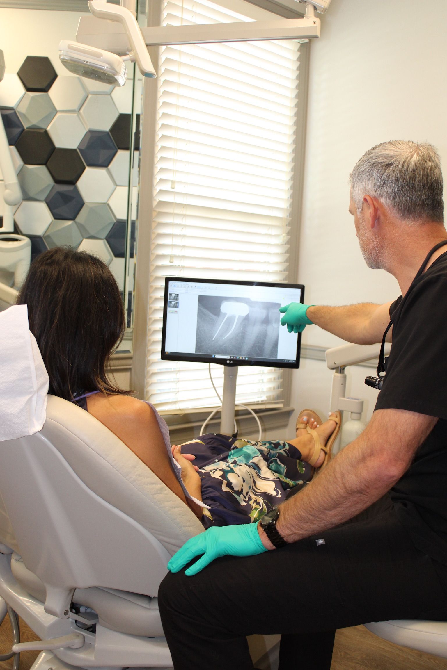 Dentist points to dental X-ray on screen, explaining it to a patient in a dental chair.