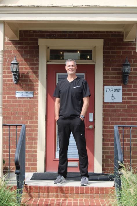 Man in black scrubs stands in front of red door of brick building; smiling, hands in pockets.
