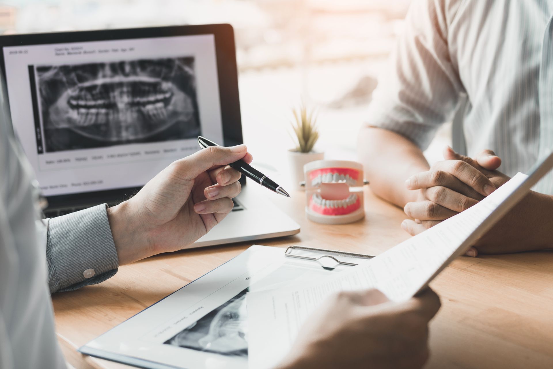 Dentist pointing at x-ray on laptop, explaining to patient. Model teeth and paperwork on the table.