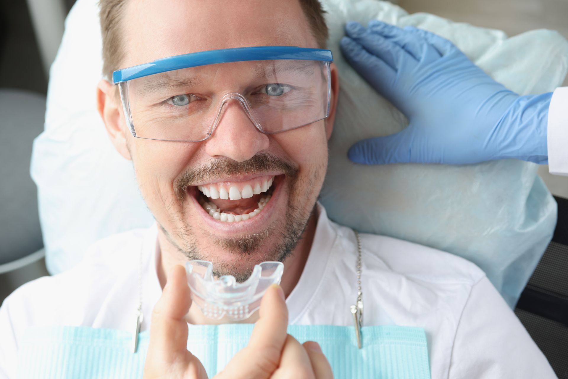 Man smiling, holding clear retainer in dental chair, wearing safety glasses; dentist's gloved hand visible.