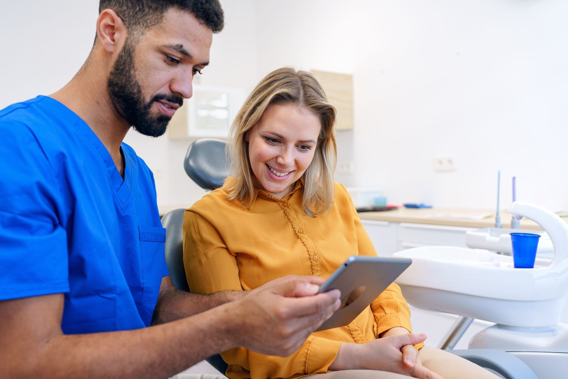 Dentist in blue scrubs shows tablet to patient in yellow shirt, both smiling in dental office.