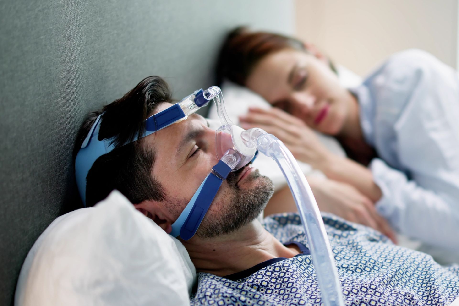 Man wearing a CPAP mask, sleeping in bed with a woman beside him.