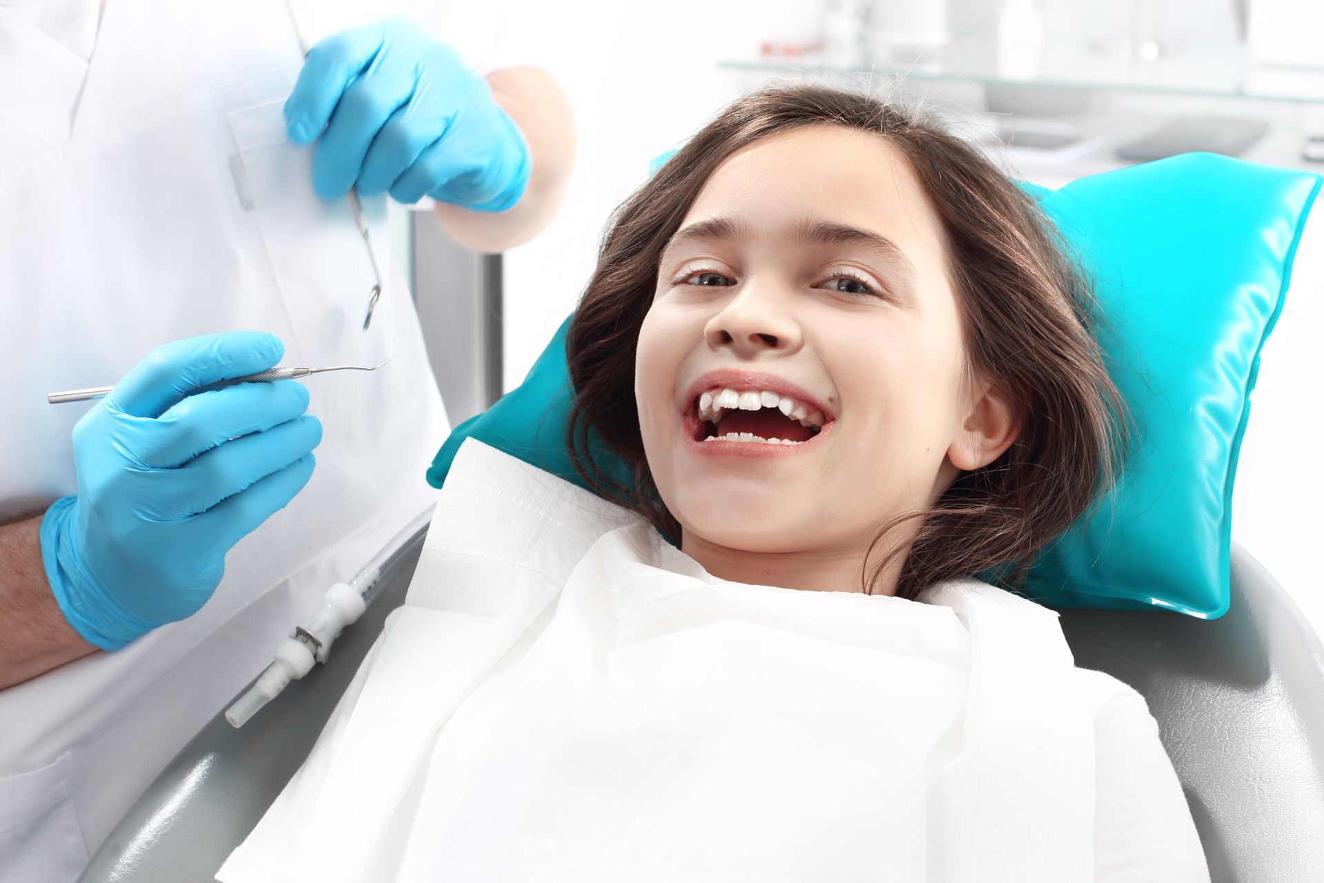 Girl smiling in a dentist chair, dentist's gloved hand holding dental tools, bright setting.