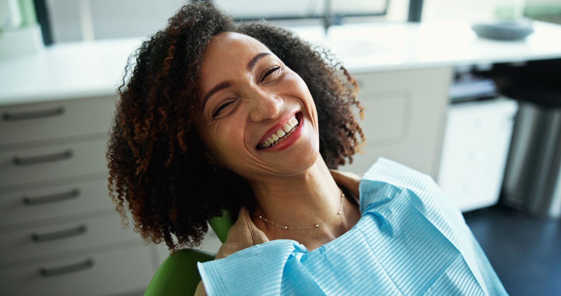 Woman with curly hair smiles broadly in a dentist's chair, wearing a blue bib.