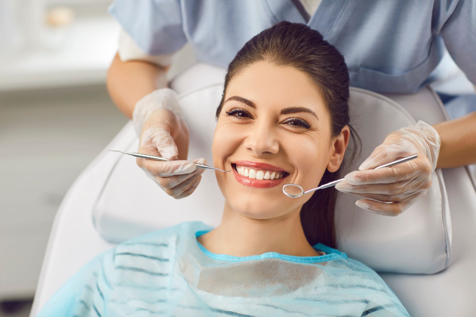 Woman smiling at dentist during a check-up. Dentist in gloves holds tools, dental chair, blue smock.